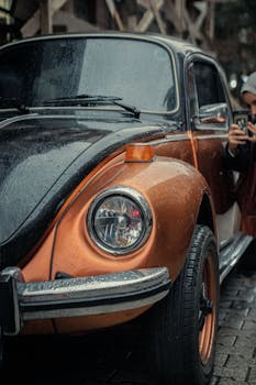 Close-up of a vintage Beetle car with rain drops, captured in an urban setting with a person in the background.