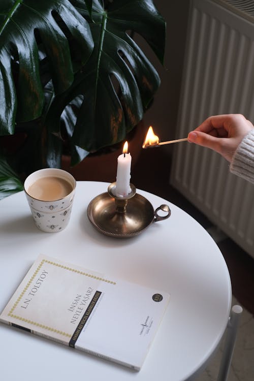 A Woman Lighting a Candle Standing on a Table next to a Book and a Cup of Coffee