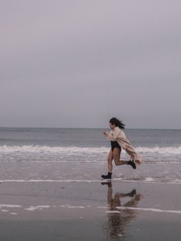 A woman runs along a cloudy beach with ocean waves and her reflection in view.