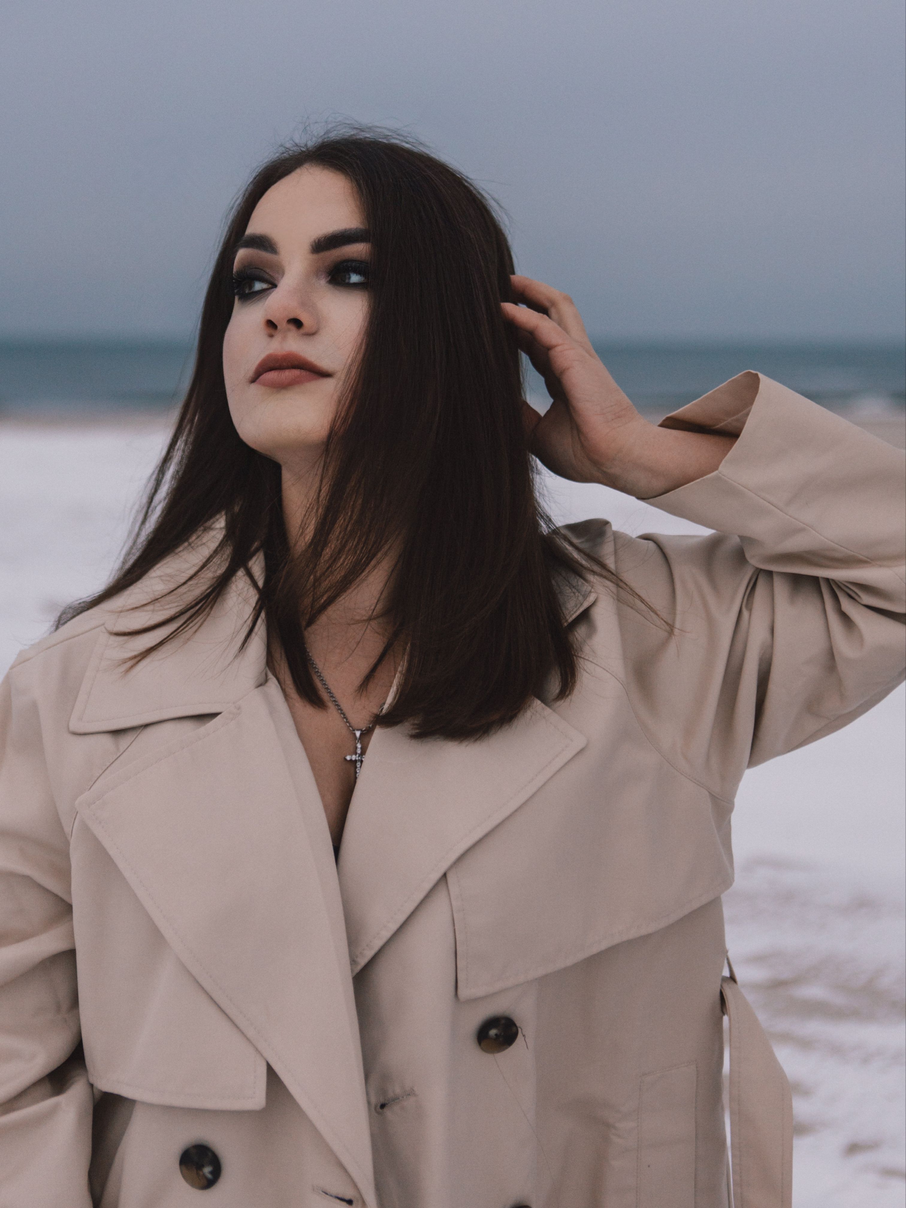 Stylish woman with black hair in a trench coat poses on a beach during winter.