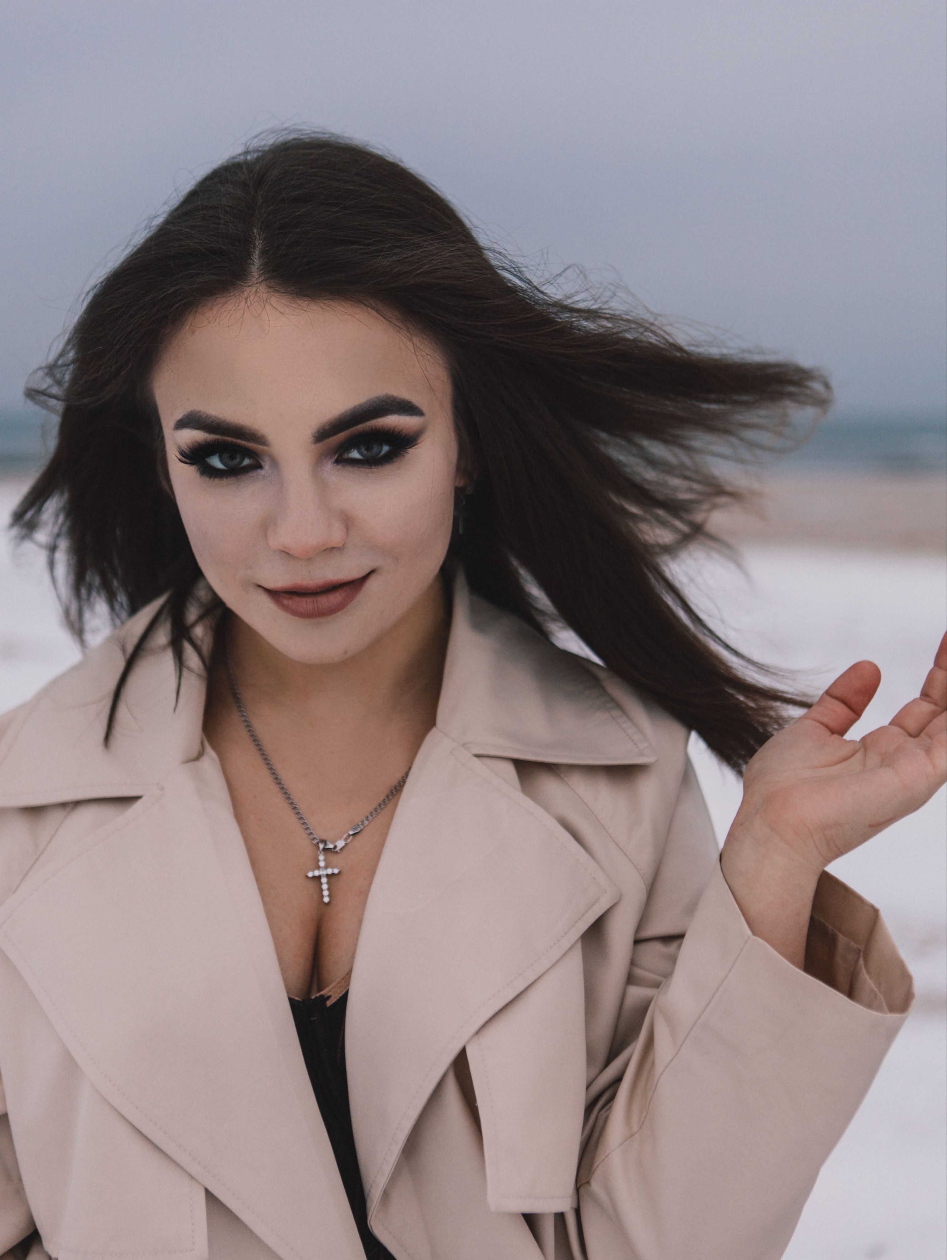 Elegant woman with dark makeup poses in a trench coat on a winter beach.