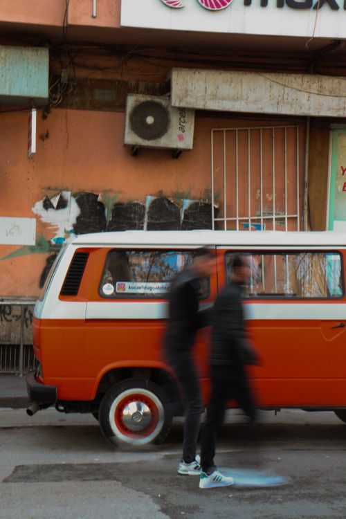 Free Street scene featuring a vintage orange van with blurred motion of two people walking. Stock Photo
