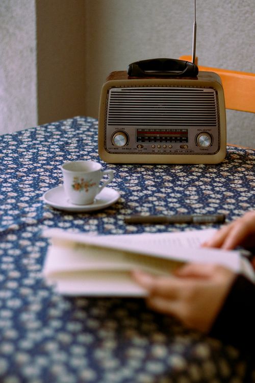 Free A cozy, vintage setting featuring a retro radio, a cup of tea, and a reader enjoying a book on a floral tablecloth. Stock Photo