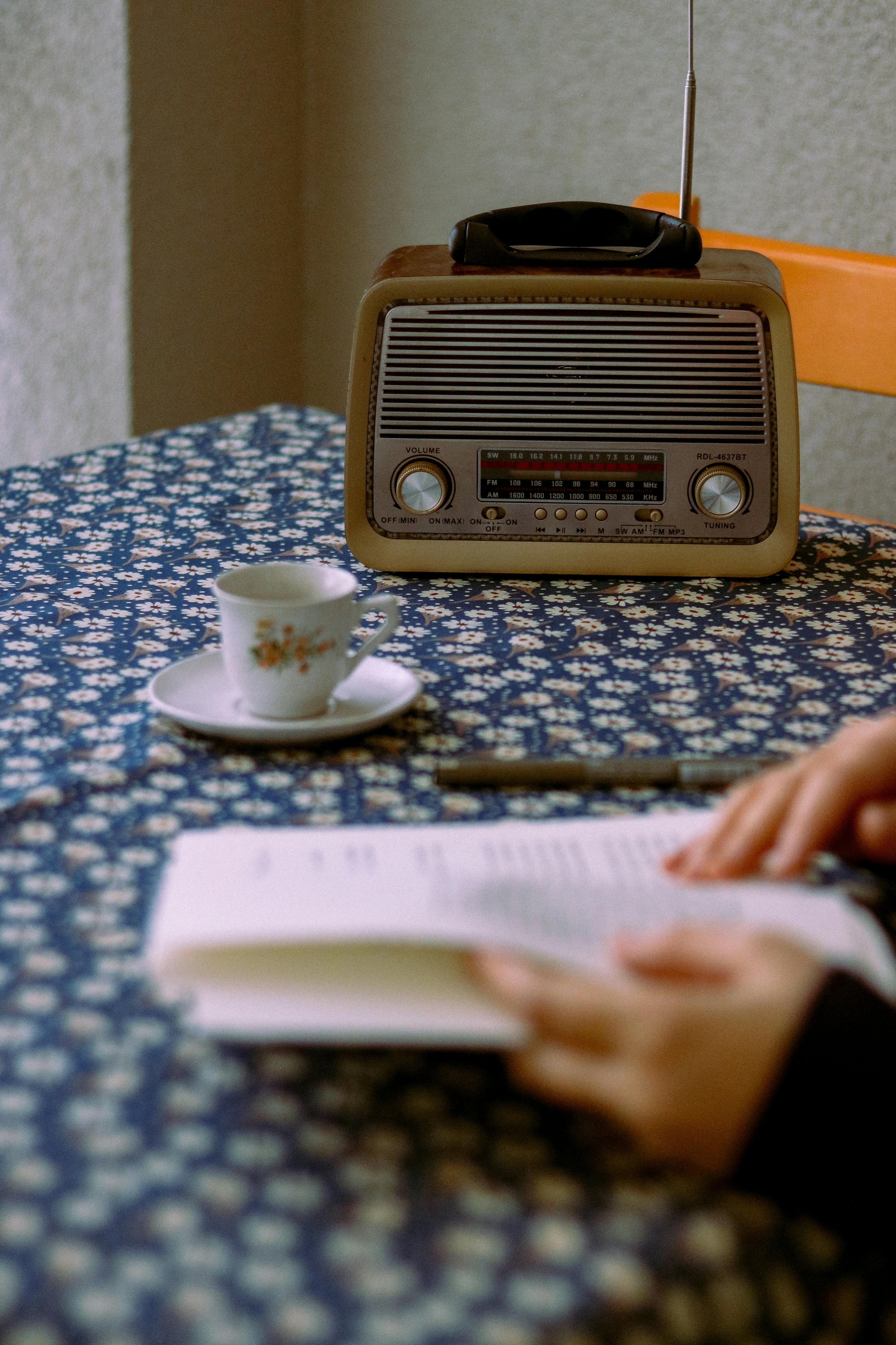 A cozy setup featuring a vintage radio, floral teacup, and book in hand on floral-patterned table.