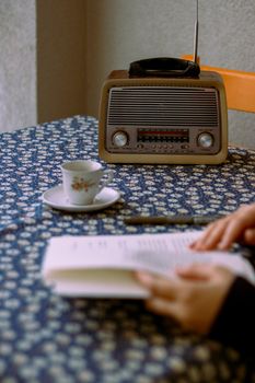 A cozy setup featuring a vintage radio, floral teacup, and book in hand on floral-patterned table.