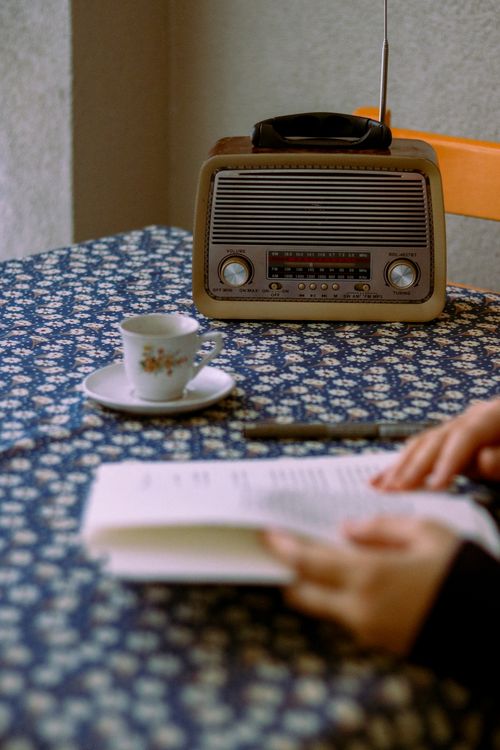 Free A cozy setup featuring a vintage radio, floral teacup, and book in hand on floral-patterned table. Stock Photo