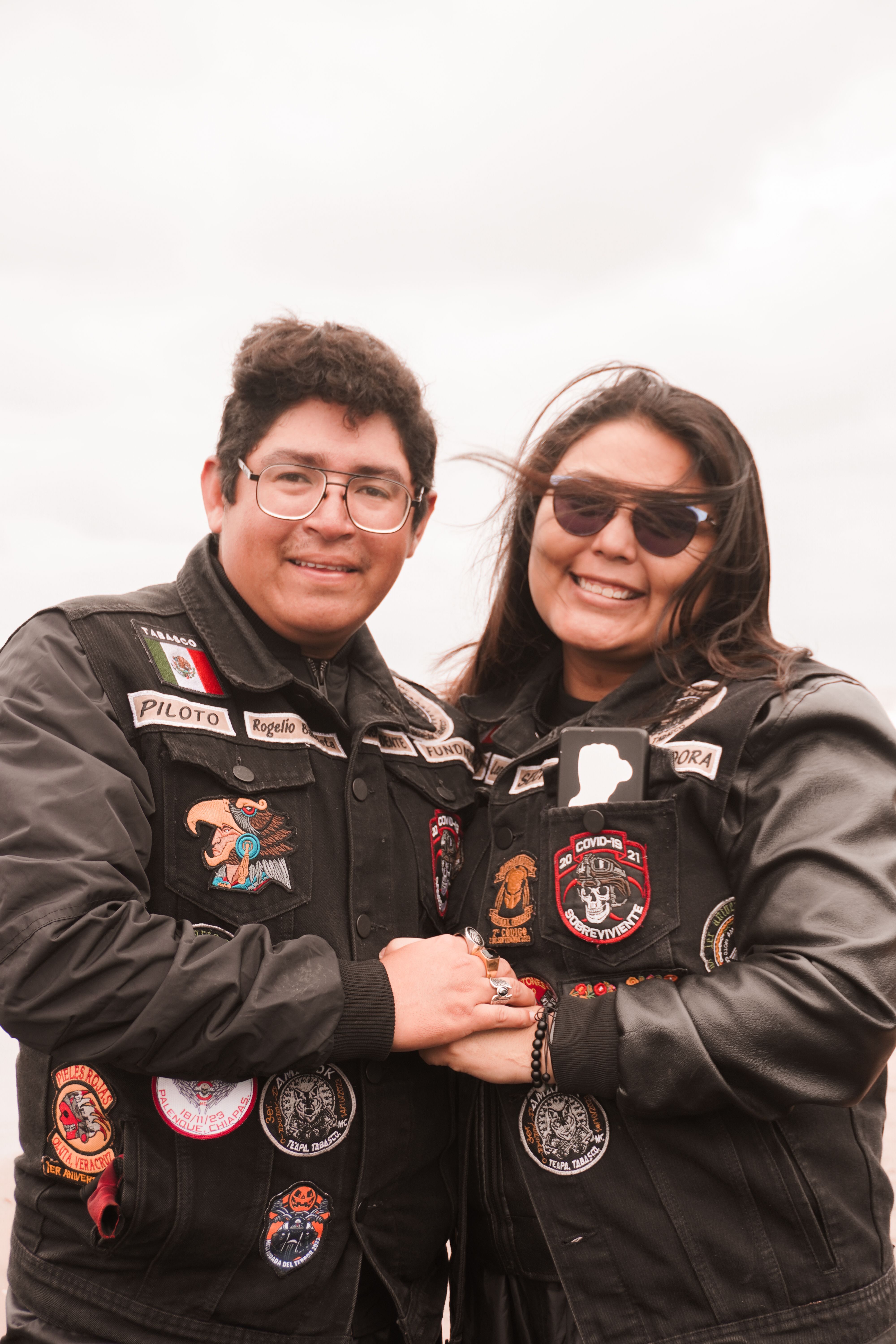 Smiling couple in biker jackets embrace outdoors, embodying happiness and unity on a cloudy day.