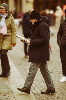 Senior man in warm jacket reads newspaper while walking on urban pavement in Padova, Italy.