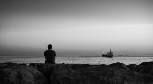 Free A person sitting on rocks by the sea, gazing at a distant ship under a moody sky. Stock Photo