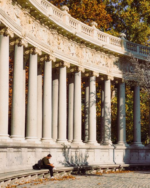A person sits by the colonnade of Alfonso XII Monument at Retiro Park, Madrid, showcasing autumn colors.