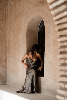Stylish woman in gray silk dress posing by an arched window in a Moroccan setting.