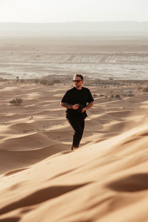 Free Man in black attire hiking on sand dunes in a desert landscape, holding a camera. Stock Photo