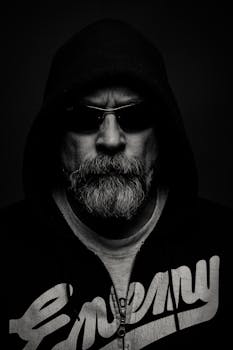 一幅戏剧性的工作室肖像，描绘了一位戴着太阳镜和连帽衫的胡须男子