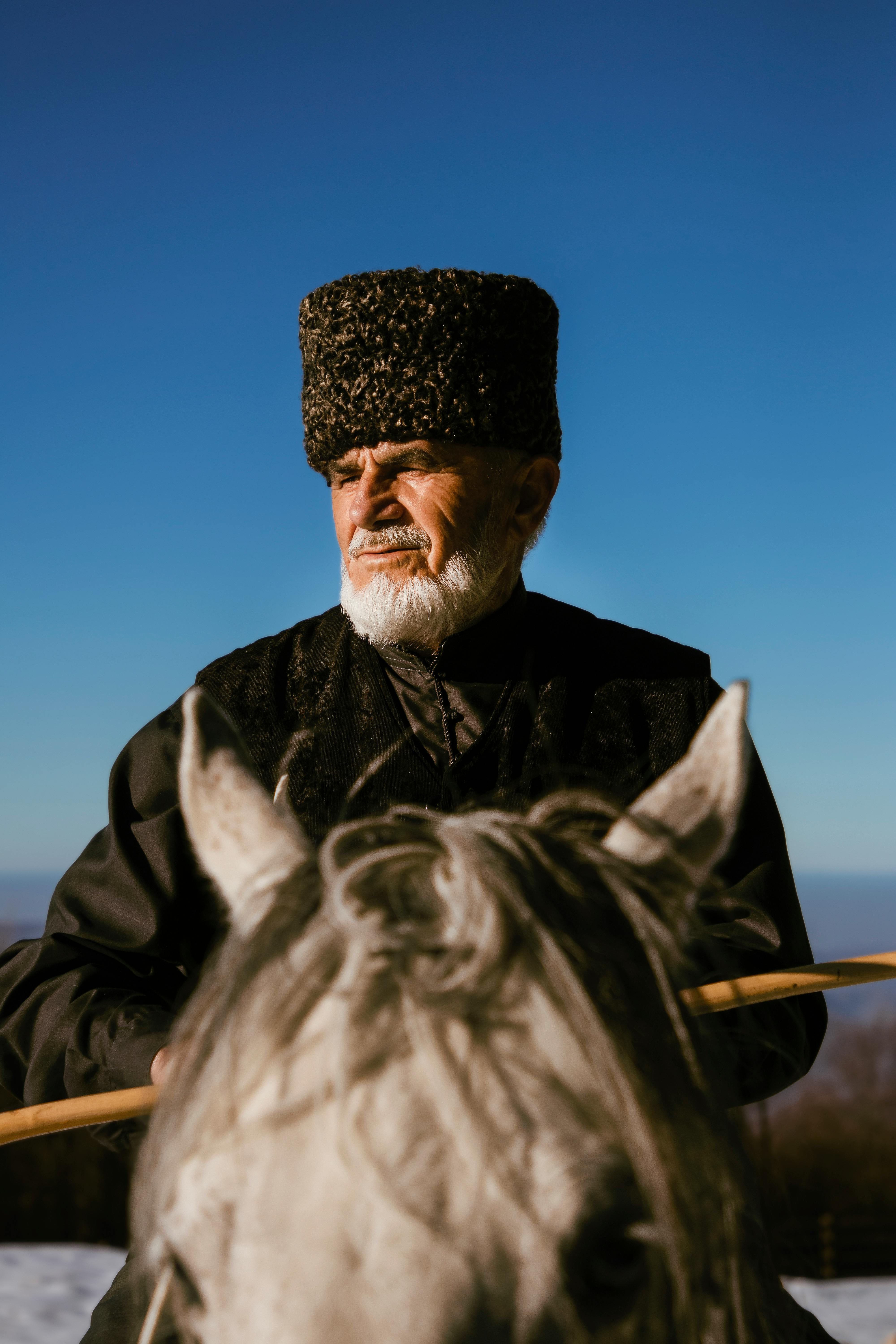 Elderly man with fur hat riding a horse in winter setting, captured in selective focus.
