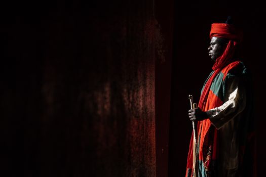 A man in traditional attire with vibrant colors standing in shadows holding a rod.