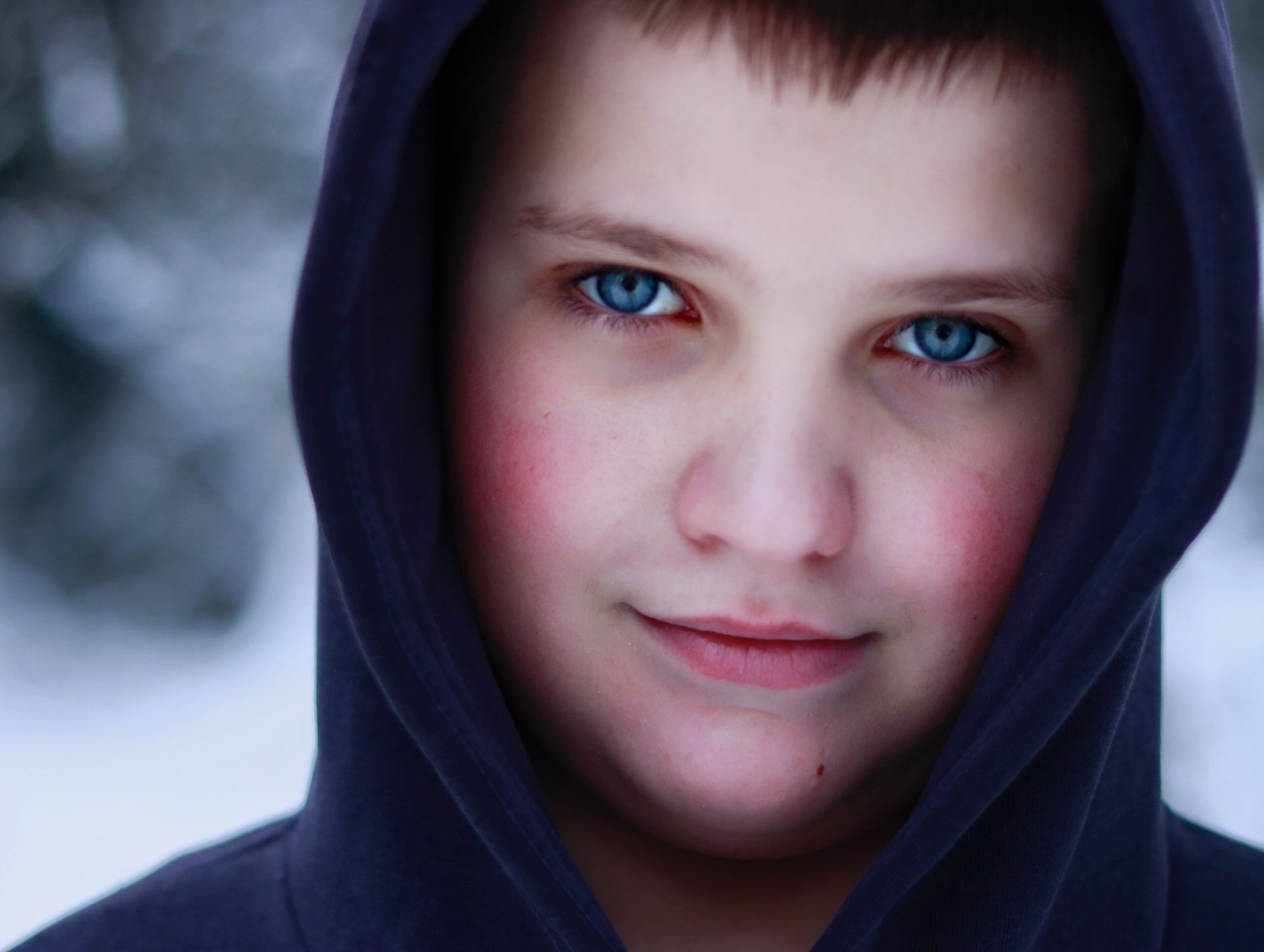 A striking close-up of a boy in a hoodie showing vivid blue eyes and rosy cheeks.