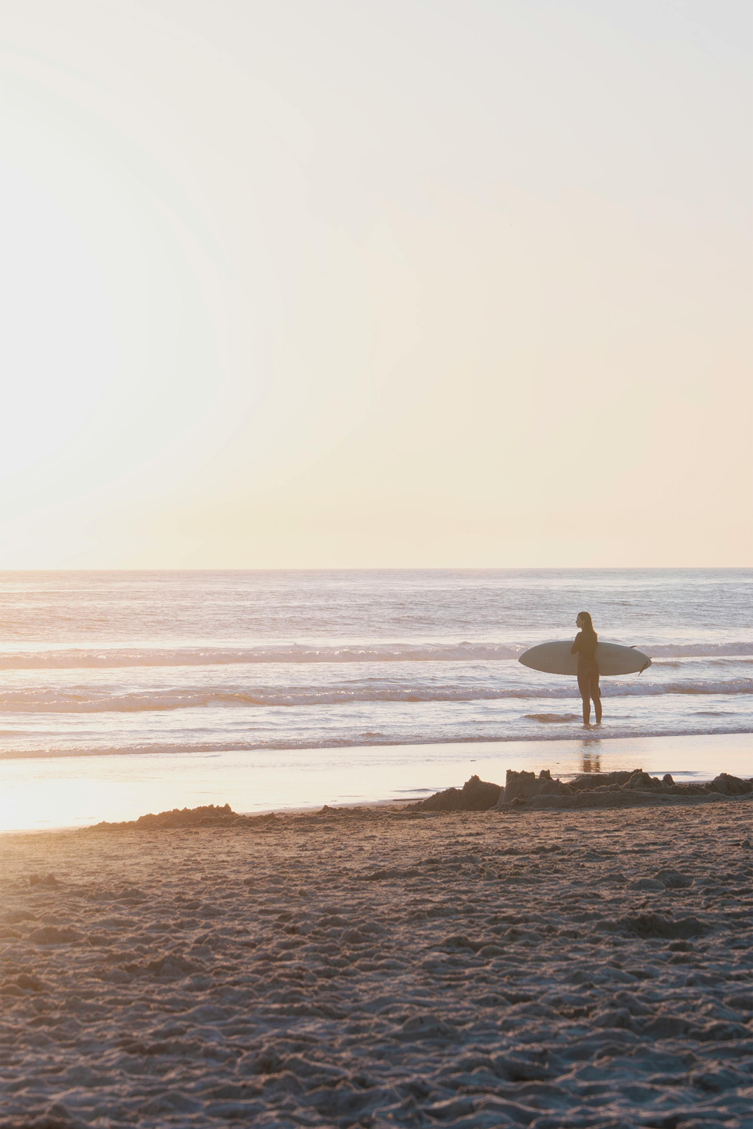 Silhouette of a surfer with a surfboard on a sandy beach during a warm sunset.