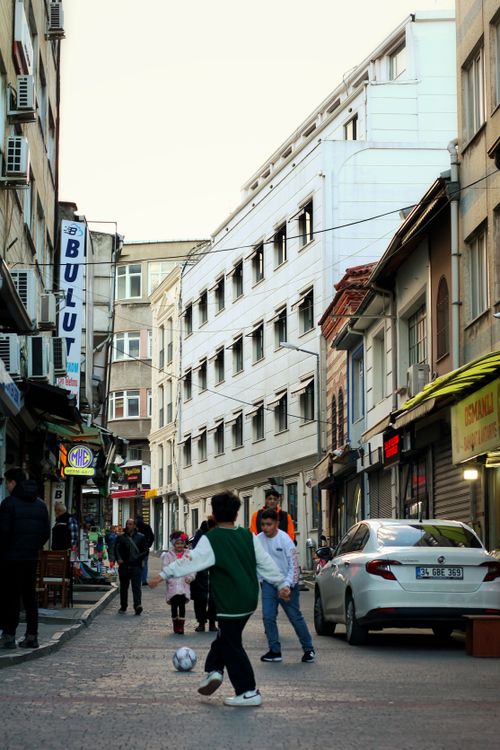 Free Kids playing soccer on a lively street in Bursa, Türkiye, surrounded by urban buildings. Stock Photo
