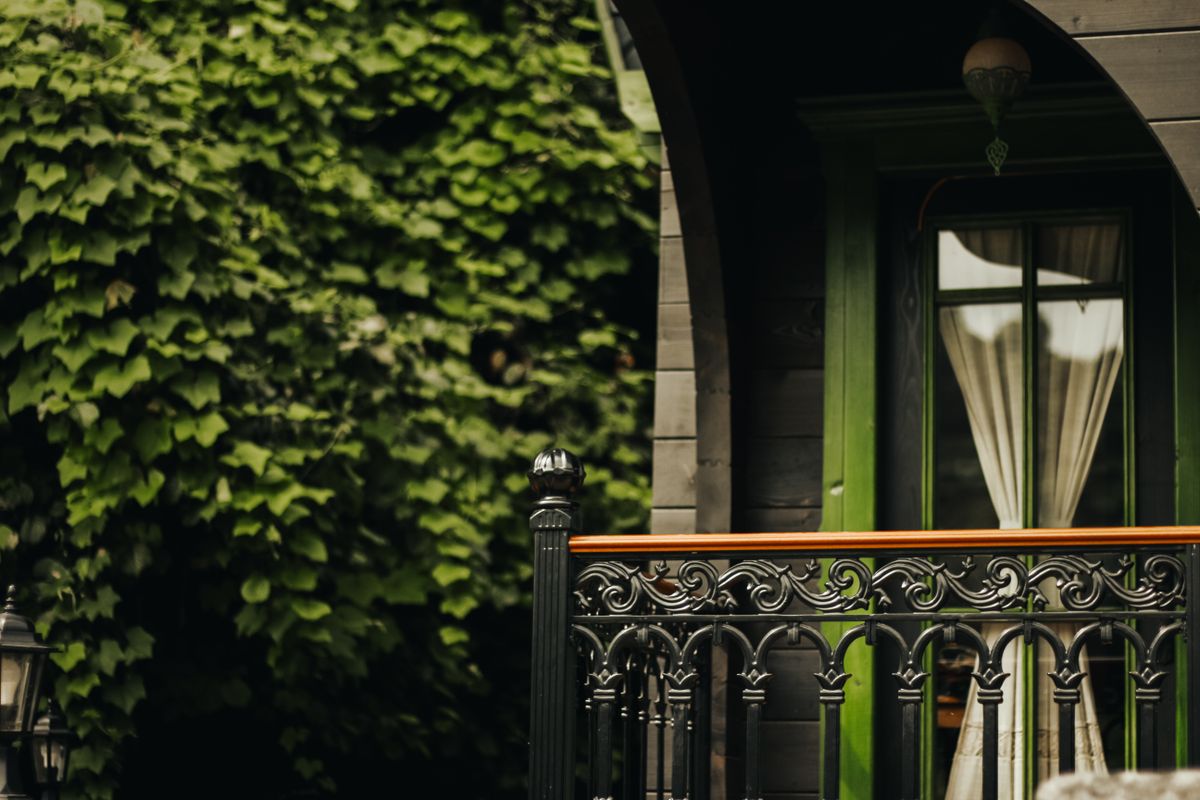 Balcon rustic cu balustradă din fier forjat cu motive ornamentale, pe fundal de vegetație bogată
