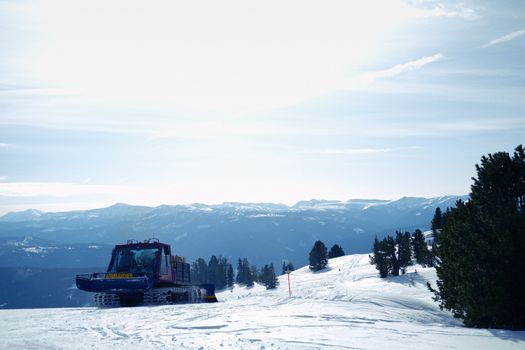 A snowcat vehicle prepares a ski slope under clear winter skies, surrounded by mountains.