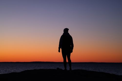 Free A solitary silhouette stands by the calm sea against a vibrant sunset sky. Stock Photo
