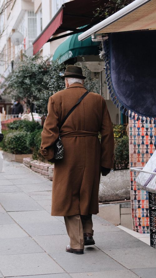 Free Elderly man in brown coat and hat walking on a city sidewalk during the day. Stock Photo