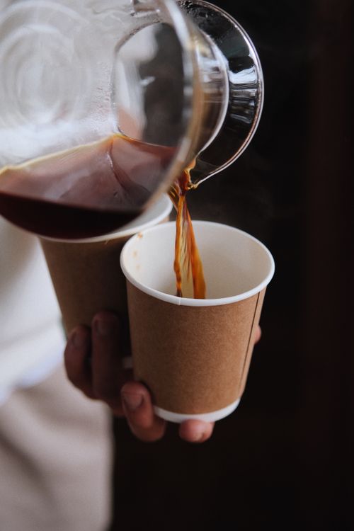 Free A hand pouring freshly brewed coffee from a glass pitcher into a paper cup indoors. Stock Photo