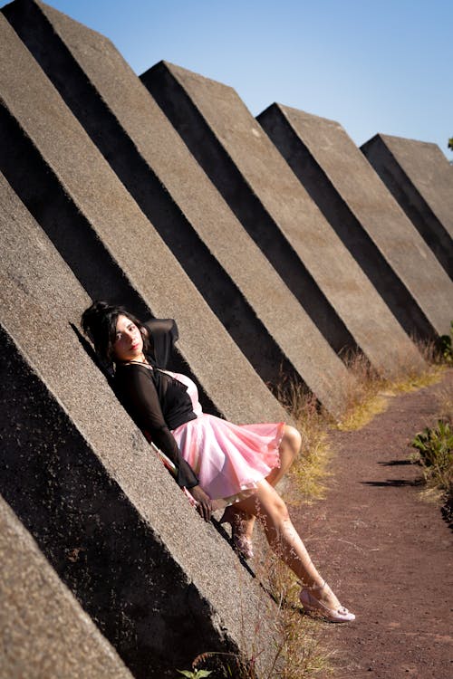A woman in a pink dress is leaning against a concrete wall