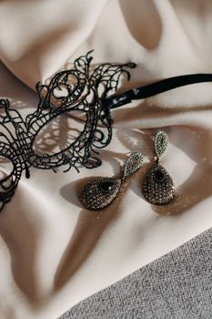 Stylish black lace mask and sparkling teardrop earrings on soft satin fabric; perfect for a masquerade theme.