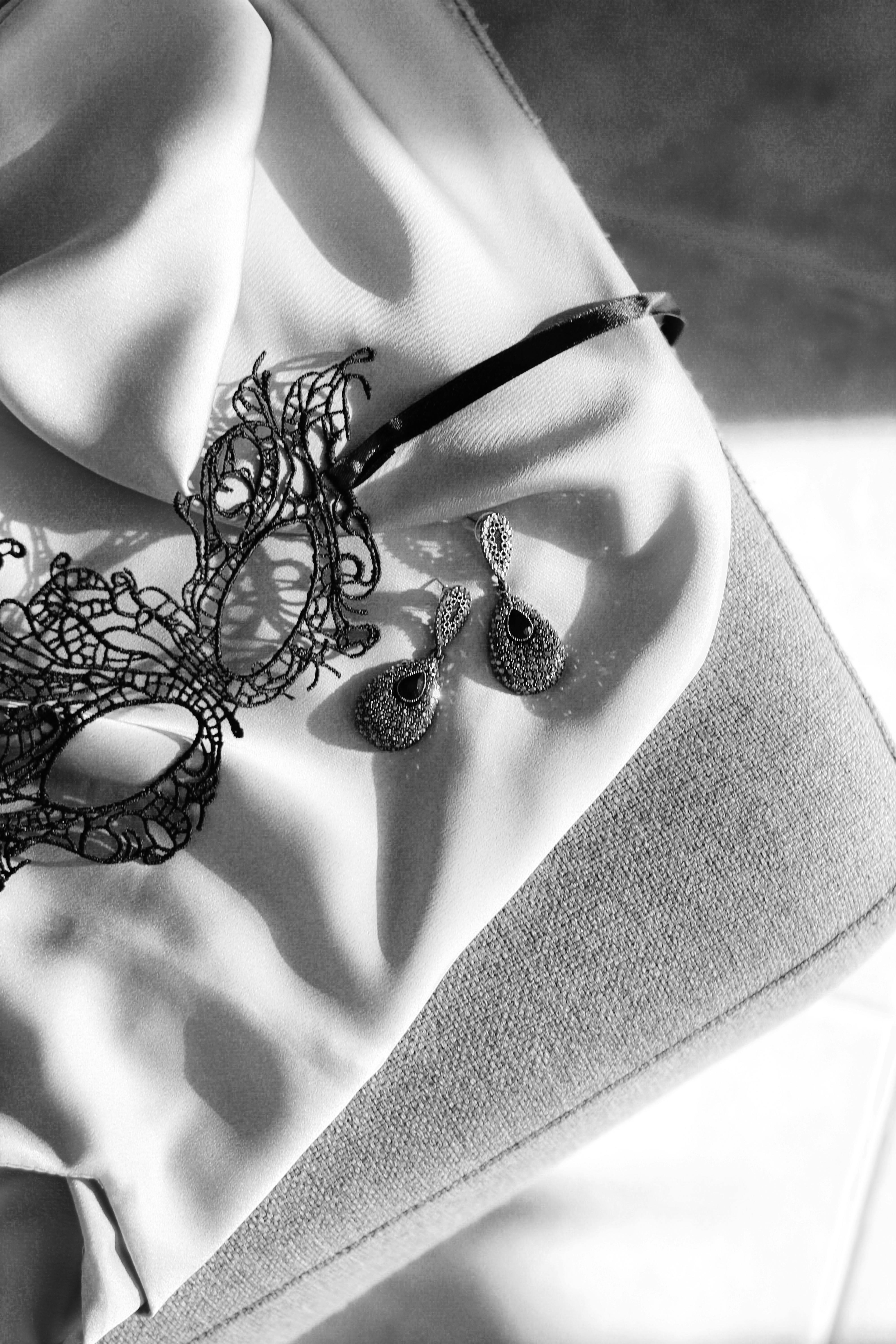 Black and white photo of a lace mask and earrings atop gently draped fabric.