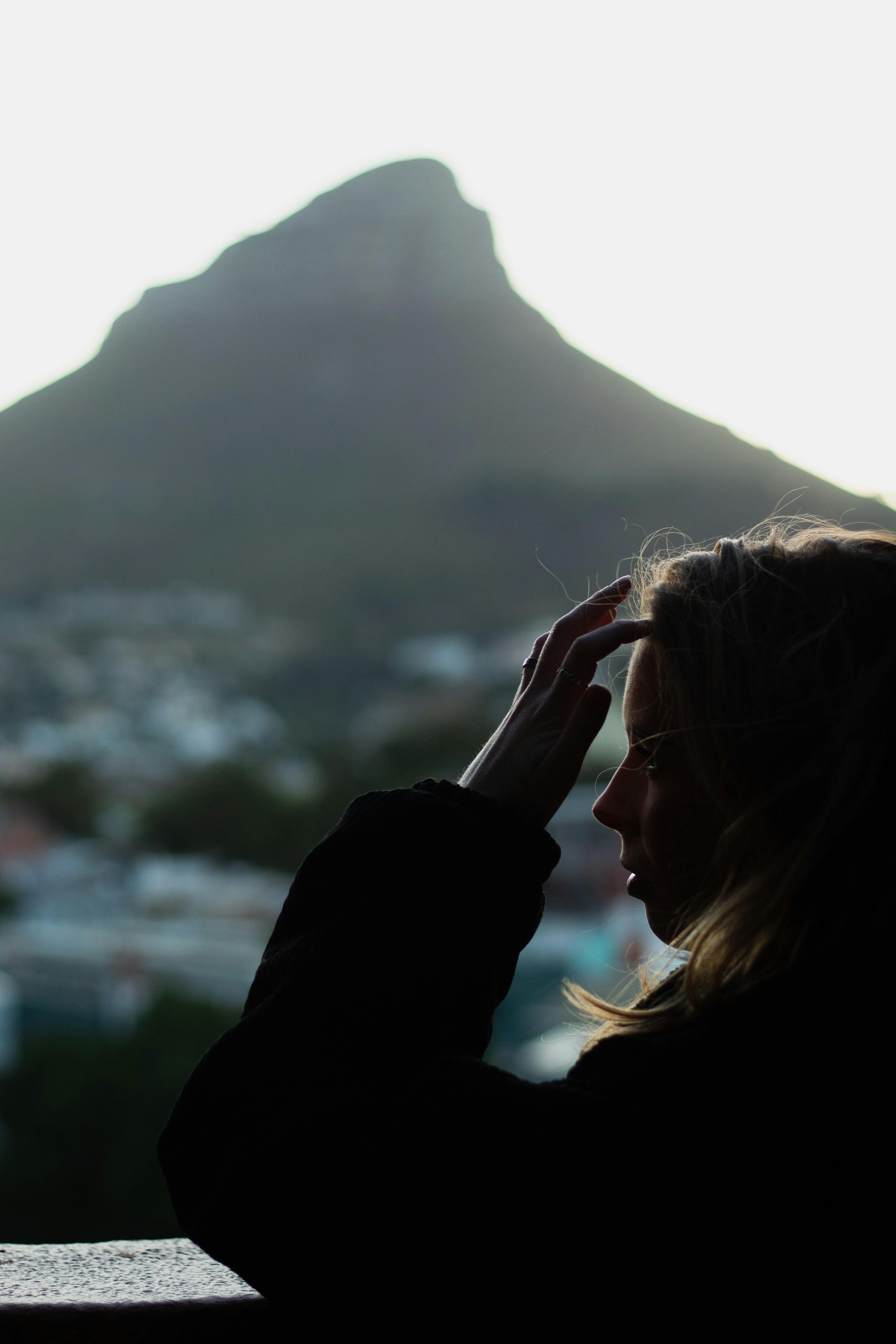 Silhouetted woman touches her hair, standing with a mountain view at sunset.