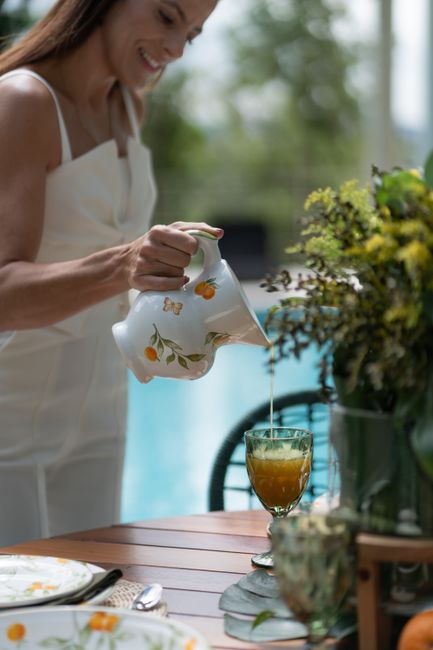 Fotomural Una mujer vierte jugo de una jarra floral en un vaso en una mesa junto a la piscina.