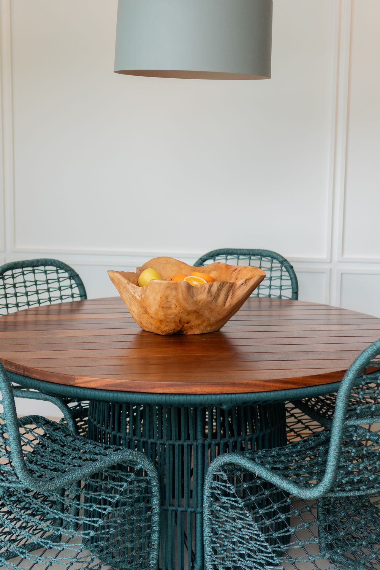 Round slatted wood table with teal woven chairs and a wood fruit bowl