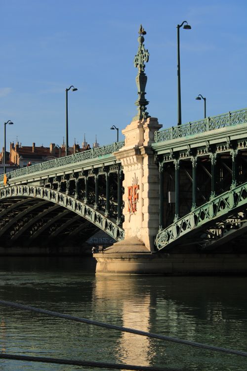 Free A majestic green metal bridge spans a calm river with urban architecture in the backdrop during sunset. Stock Photo