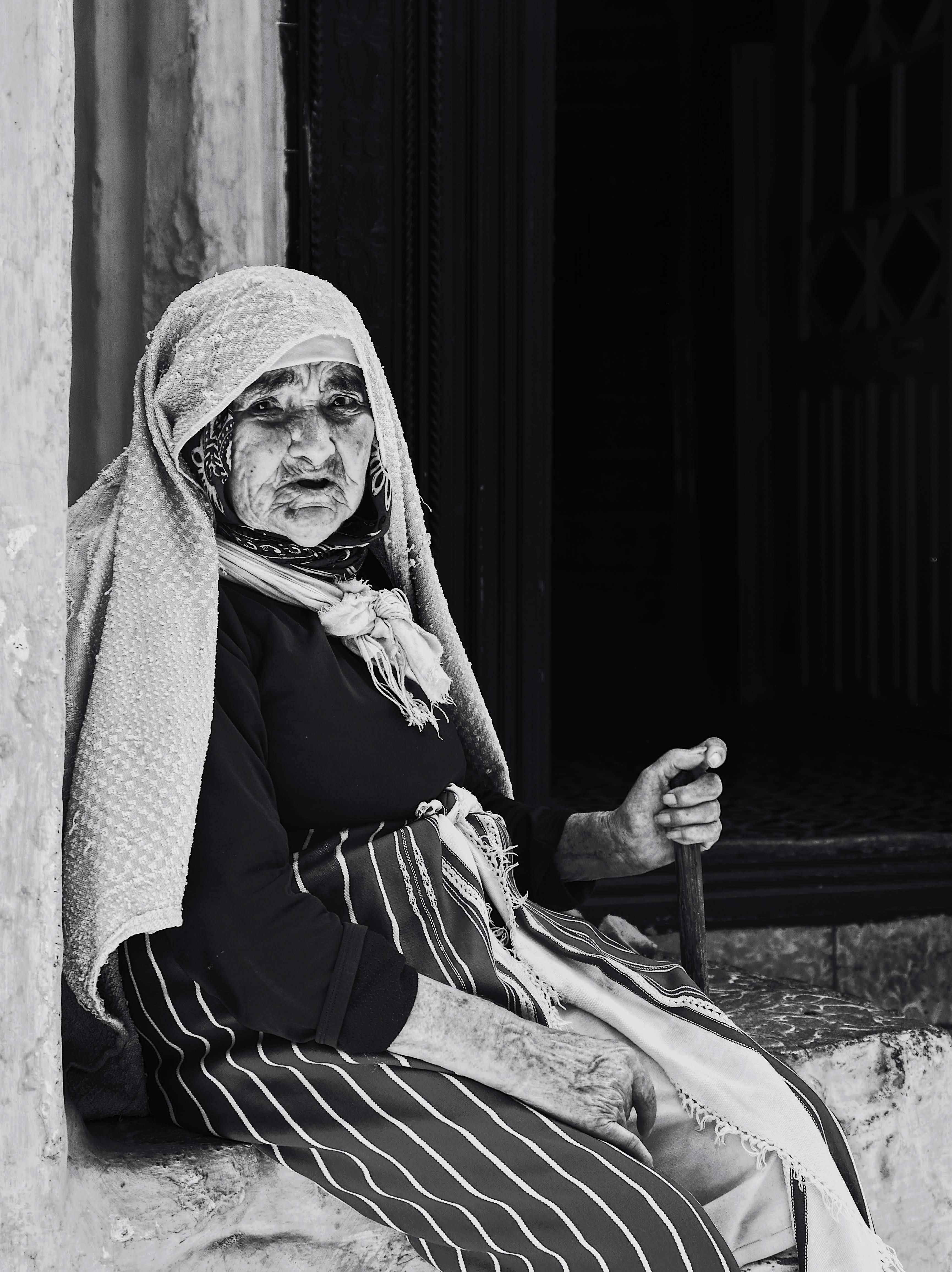 Black and white photo of an elderly woman sitting by a window holding a stick in a rural setting.