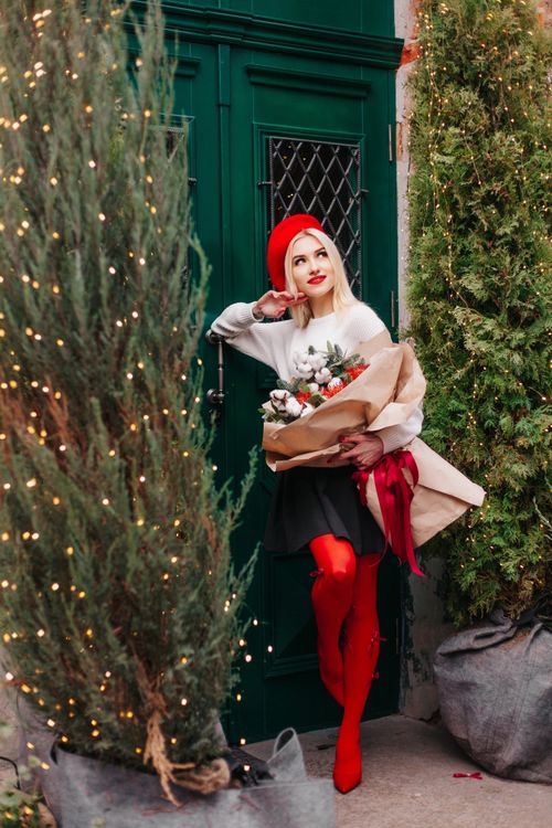 Free Young woman in festive attire holding bouquet amidst Christmas decorations and urban greenery. Stock Photo