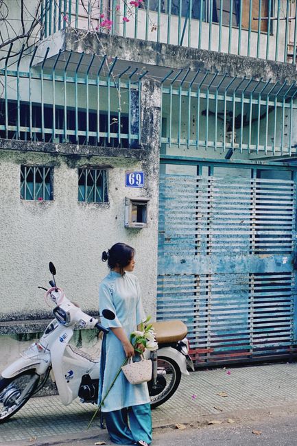Vietnamese woman in traditional ao dai standing by a motorbike in an urban city setting.