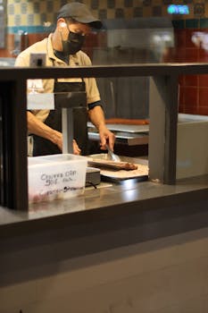 A chef wearing a mask slices food inside a restaurant kitchen during the day.