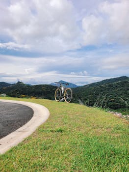 A lone bicycle rests on a grassy hill beside a winding road under a cloudy sky.