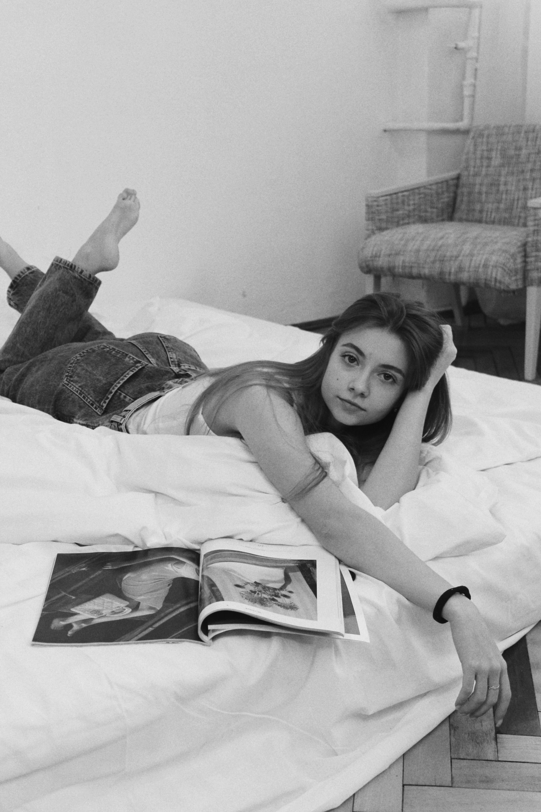 Young woman relaxing barefoot on bed with a magazine in a casual indoor setting.