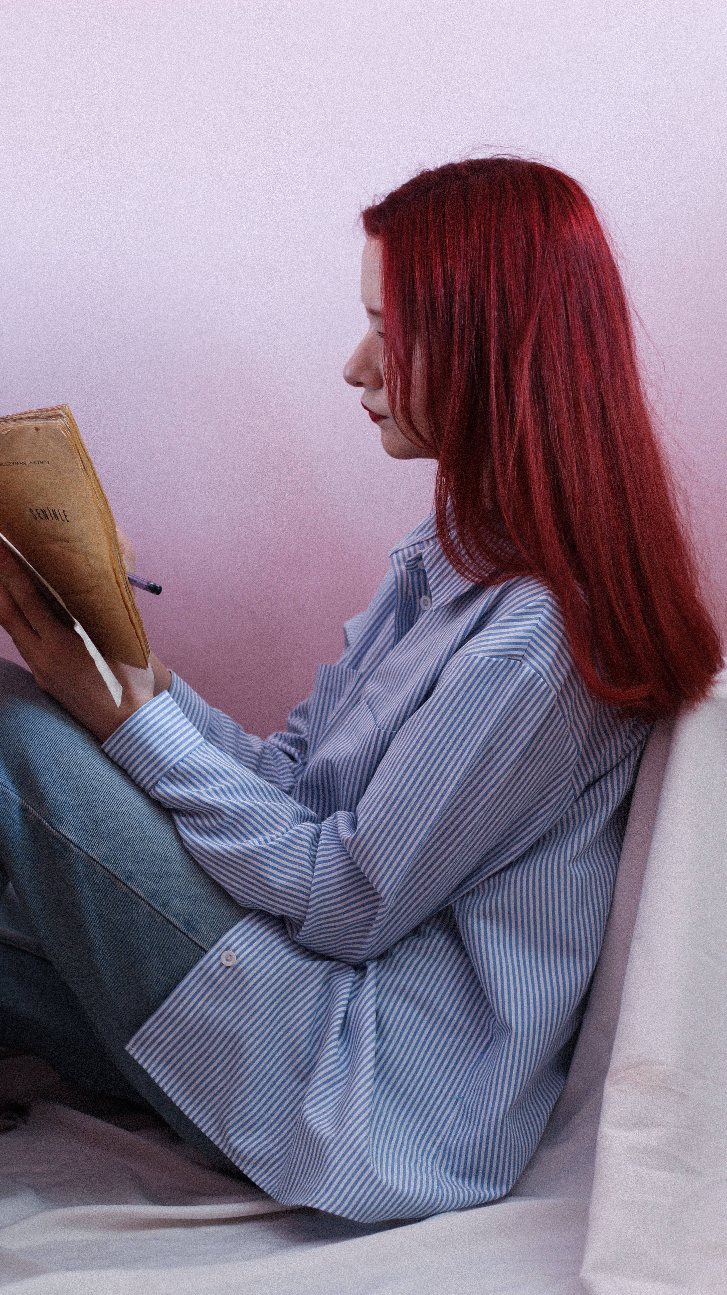 Caucasian redhead woman reading book in cozy setting, wearing striped shirt.
