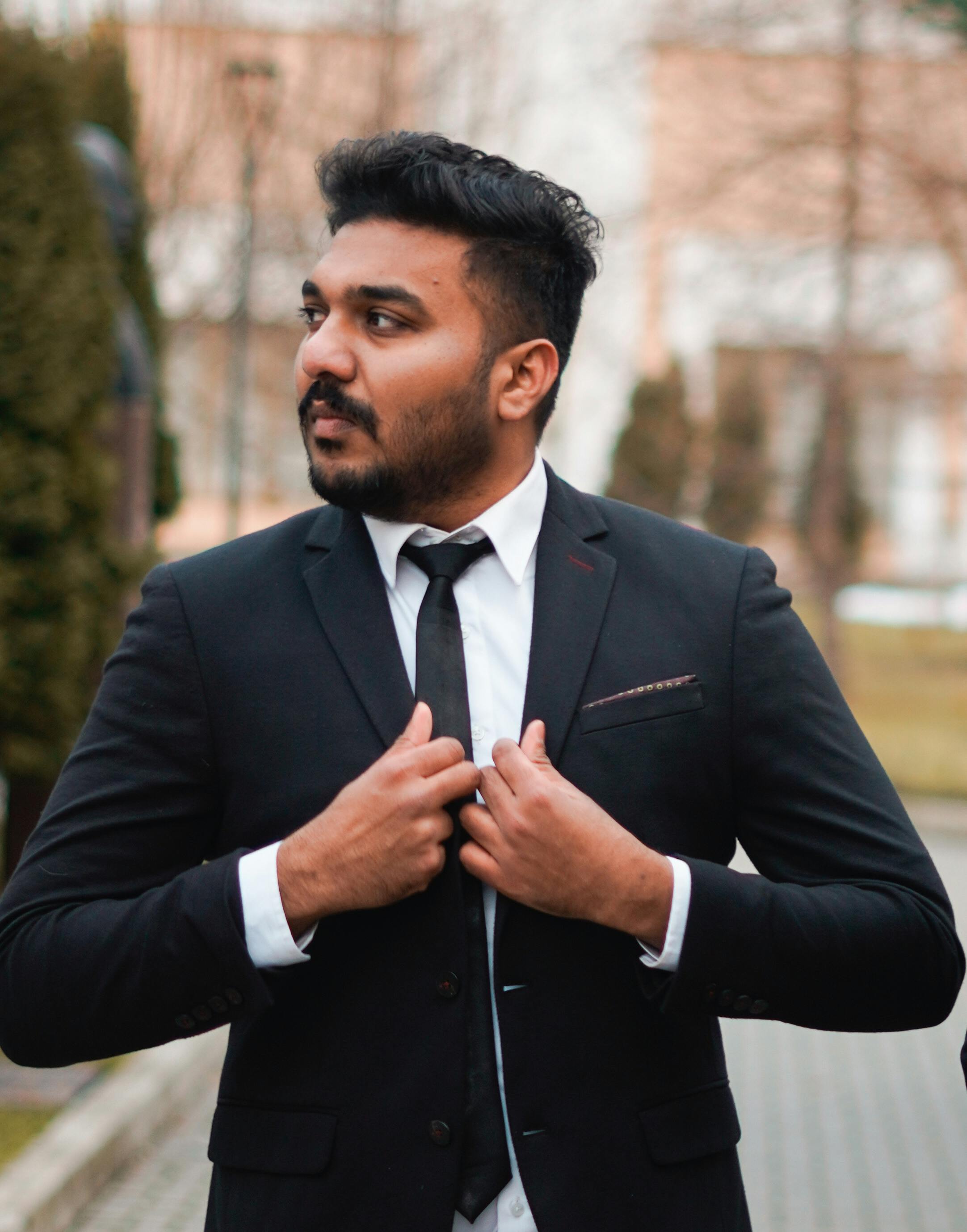 Young man in a black suit adjusting his tie outdoors, exuding confidence and elegance.