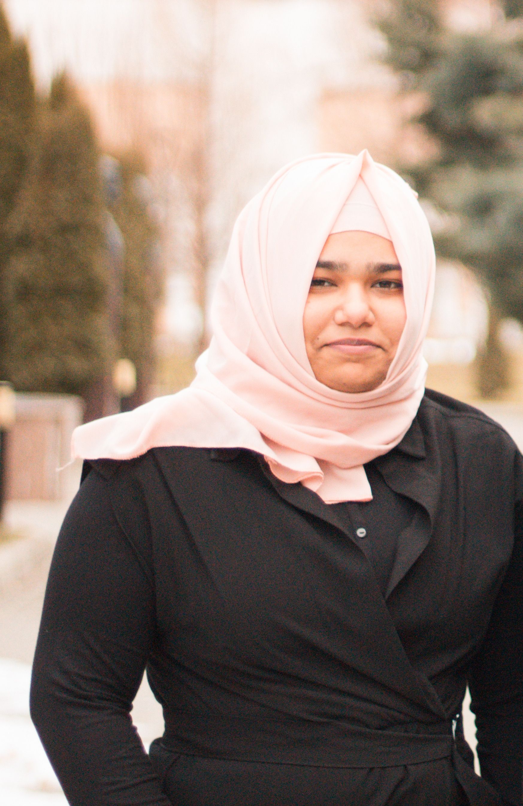 A serene portrait of a smiling woman wearing a pink hijab and black dress in an outdoor setting.