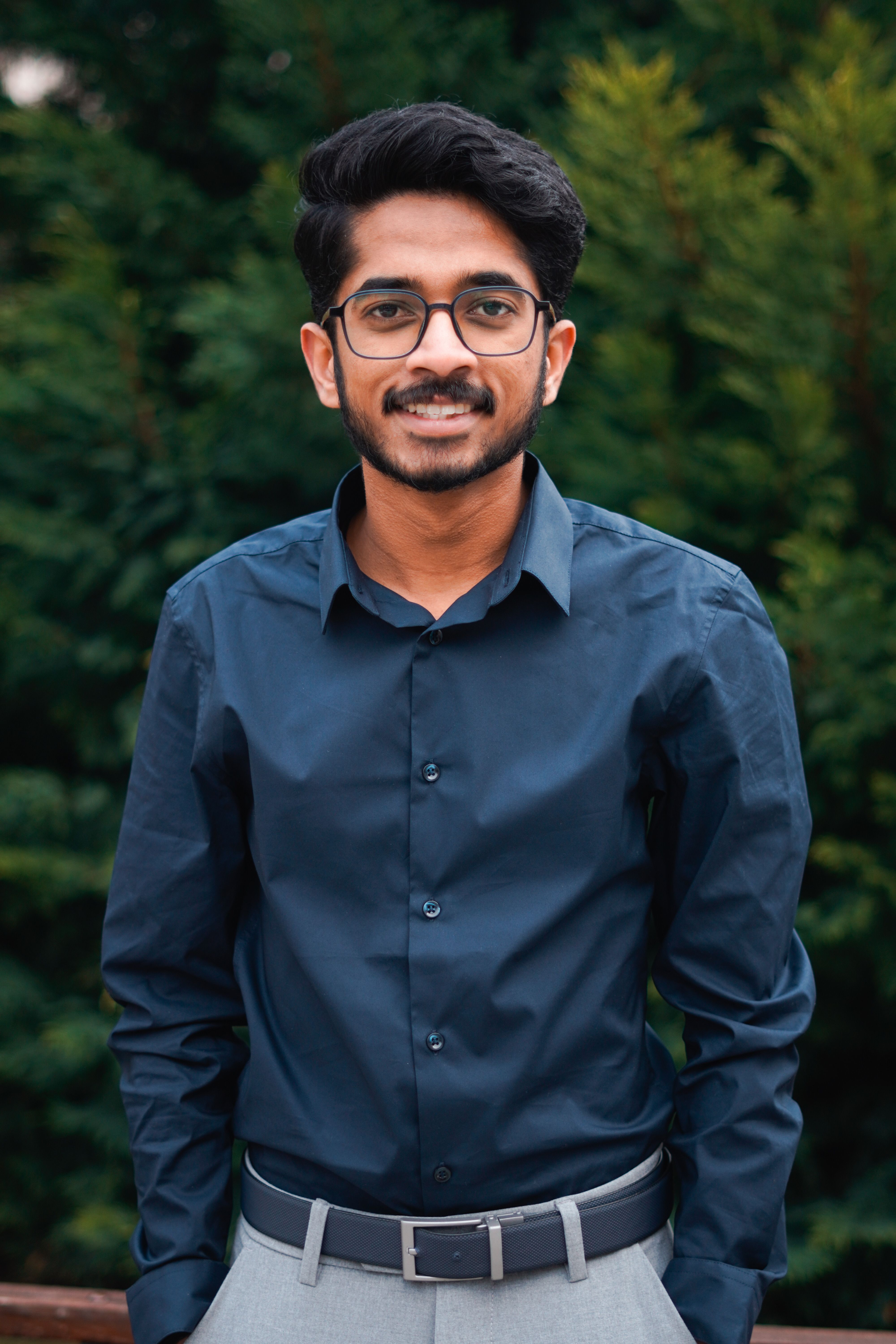 Portrait of a young man with glasses and mustache smiling outdoors with greenery background.