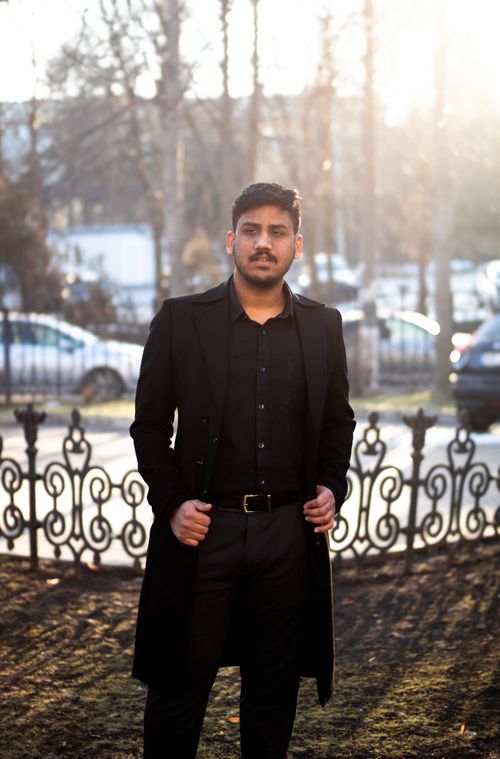 Free Young man in a long black coat standing outdoors with sunlight filtering through trees. Stock Photo