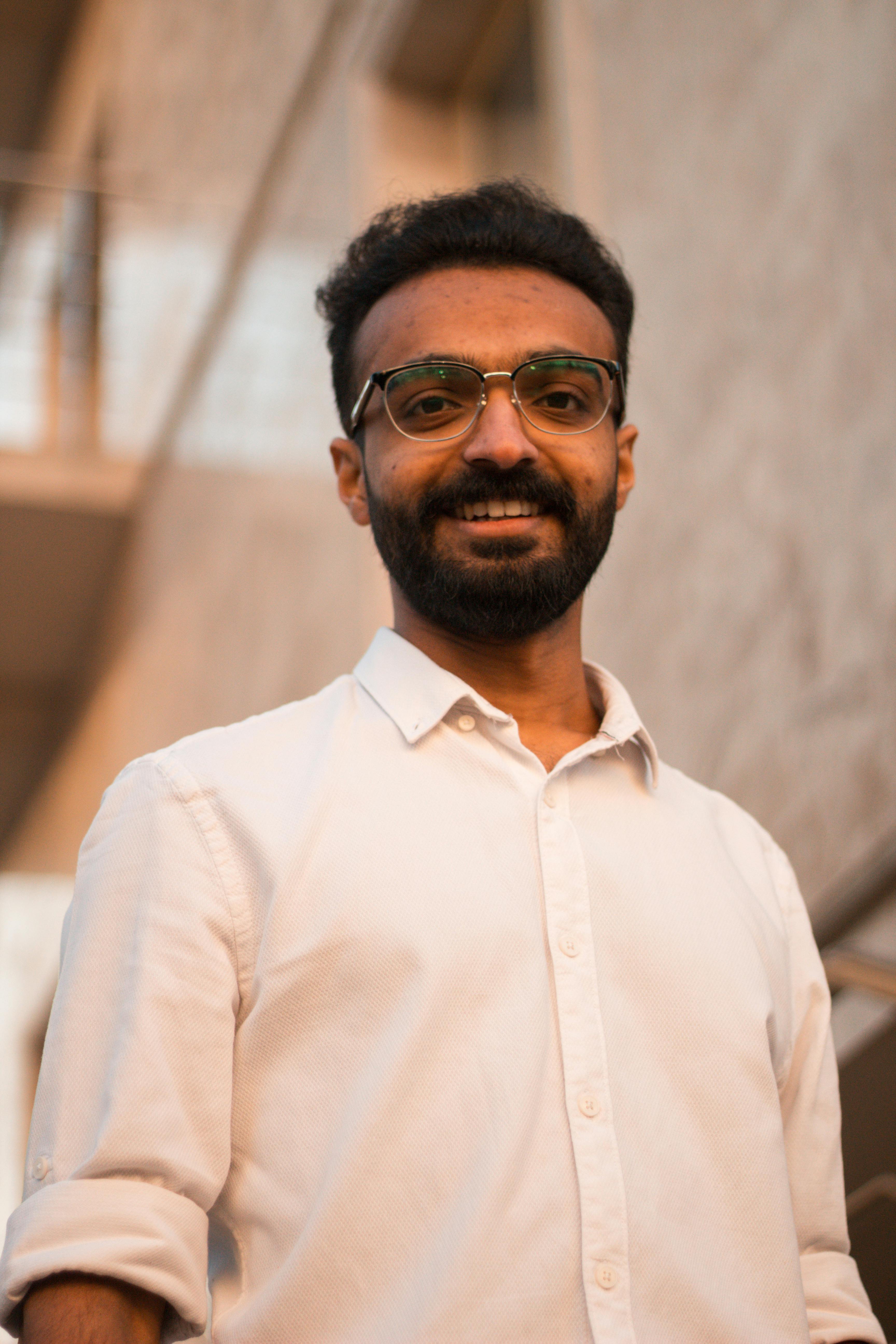 Portrait of a smiling young man with eyeglasses and beard, wearing a white shirt and standing outdoors in warm light.