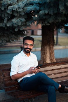 Smiling man in a white shirt sitting on a bench under a tree outdoors.