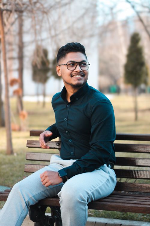 Free Stylish man in glasses smiling while seated on a park bench amidst trees on a sunny day. Stock Photo