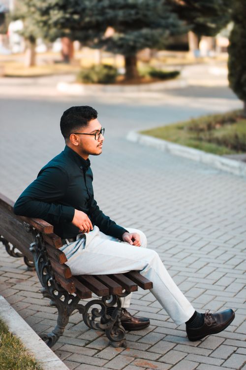 Free Young man sitting elegantly on a park bench, dressed in casual fashion with eyeglasses. Stock Photo