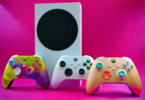 Vibrant gaming controllers displayed with a console on a bold pink backdrop.
